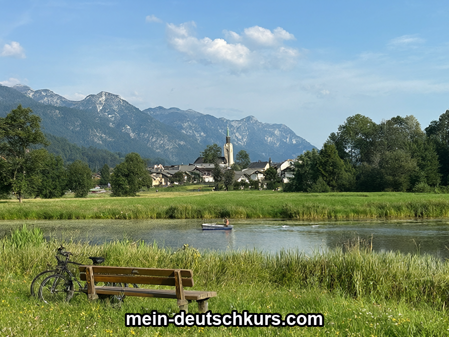 Eine Landschaft mit Bank und Fahrrad auf einer Wiese, einem kleinen See mit Boot, einem Dorf mit Kirchturm und Bergen